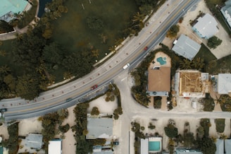 An aerial view of a suburban area featuring a curving road with several cars, surrounded by residential houses and lush greenery. One house has an unfinished roof, indicating construction work, while another has a visible swimming pool in the backyard. The arrangement of houses and roads is complemented by the presence of palm trees and other vegetation.