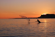 A silhouette of a person casting a fishing net into the water during sunset. The sky is painted in shades of orange and yellow as the sun sets near a distant building along the shoreline.