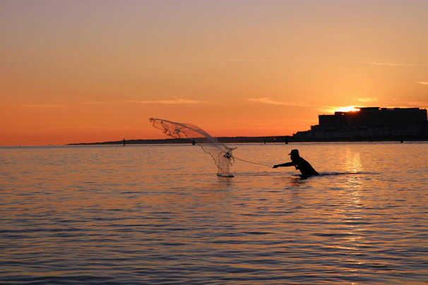 A fisherman casting his net into the river, silhouetted against a glowing sunset.