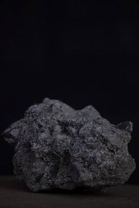 A rough, textured rock with a jagged, uneven surface sits against a dark background. The rock appears to be made of a coarse, granular material with variations in shading that suggest different mineral compositions.