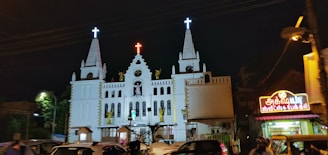 A large white church is brightly lit in the night, featuring multiple spires topped with crosses. Statues are prominently displayed on the facade, and a vibrant red neon cross is situated at the peak of the central tower. To the right, there is a well-lit shop with a colorful sign written in Tamil script. Several vehicles and people are present in the foreground, suggesting a busy urban setting.