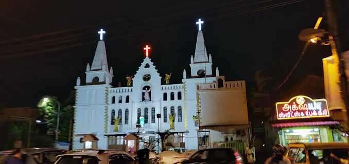 A large white church is brightly lit in the night, featuring multiple spires topped with crosses. Statues are prominently displayed on the facade, and a vibrant red neon cross is situated at the peak of the central tower. To the right, there is a well-lit shop with a colorful sign written in Tamil script. Several vehicles and people are present in the foreground, suggesting a busy urban setting.