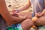 Two people are seated cross-legged on a sandy beach, engaged in meditation or a mindful practice. Their hands are resting on their knees in a meditative gesture, and they are partially visible from the shoulders down. The beach surface is scattered with small rocks and debris.