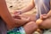 Two people are seated cross-legged on a sandy beach, engaged in meditation or a mindful practice. Their hands are resting on their knees in a meditative gesture, and they are partially visible from the shoulders down. The beach surface is scattered with small rocks and debris.