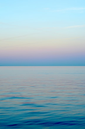 A serene seascape with a calm ocean and a horizon blending into the sky. The sky transitions from a bright blue at the top to a gentle gradient of pastel hues closer to the horizon.