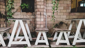 A rustic outdoor setting features wooden tables and benches with white bases set against a brick wall. Green vines hang down the wall, adding a touch of nature to the scene. The wooden surfaces have a weathered appearance, enhancing the rustic charm.