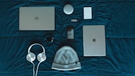On a dark teal surface, a variety of items are neatly arranged. The items include a Dell laptop, a silver iPad, a white beanie hat, white headphones, a smartphone with a dark case, a black fitness tracker, a white cylindrical speaker, a white external hard drive, and a small USB drive.
