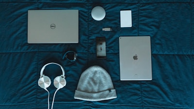 On a dark teal surface, a variety of items are neatly arranged. The items include a Dell laptop, a silver iPad, a white beanie hat, white headphones, a smartphone with a dark case, a black fitness tracker, a white cylindrical speaker, a white external hard drive, and a small USB drive.