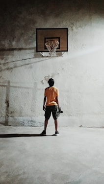 A person stands alone on a concrete floor, facing a basketball hoop mounted on a worn wall. The lighting casts a long shadow and highlights the texture of the wall. The individual appears to be holding a basketball.