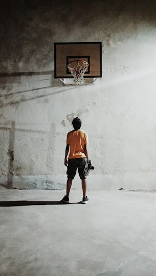 A person stands alone on a concrete floor, facing a basketball hoop mounted on a worn wall. The lighting casts a long shadow and highlights the texture of the wall. The individual appears to be holding a basketball.