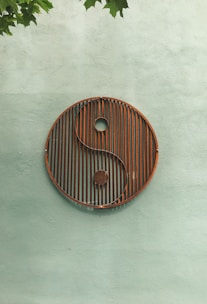 A large yin-yang symbol made of metal with rust accents is mounted on a textured light green wall. Green leaves extend from the upper left corner, adding a natural contrast to the geometric simplicity of the symbol.