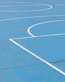 A bright blue outdoor basketball court with white lines marking various sections such as the three-point line and key area. The surface appears smooth and well-maintained.