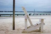A white wooden Adirondack chair is positioned on a sandy beach with a calm body of water in the background. The sky is overcast, creating a subdued atmosphere. A metallic pole stands near the chair, and a railing separates the sand from the water.