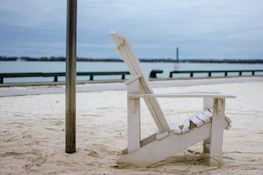 A white wooden Adirondack chair is positioned on a sandy beach with a calm body of water in the background. The sky is overcast, creating a subdued atmosphere. A metallic pole stands near the chair, and a railing separates the sand from the water.