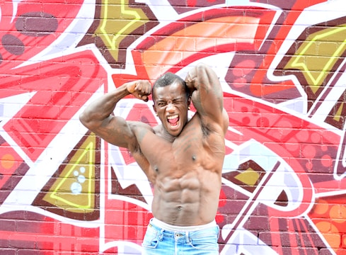 A muscular man flexes his biceps and poses energetically against a vibrant graffiti-covered wall. The wall features bold abstract designs with various geometric shapes and colors, creating a lively and dynamic background.