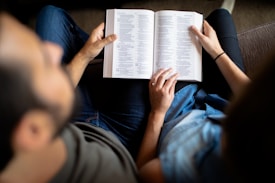 Two people are sitting closely together, holding open a book. The focus is primarily on the book, which is centered between them. They appear to be reading, with their heads looking downward towards the text. One person is wearing a blue shirt, while the other has a darker top. Light and shadows play across the scene, highlighting the intimacy of the moment.