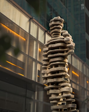 A modern sculpture composed of stacked, abstract, wavy stone slabs creating a human-like form. The sculpture is set against a backdrop of a glass and metal building with warm interior lights visible through the windows.