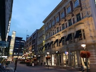 A bustling urban street scene with multi-story buildings lining both sides. The architecture is a mix of modern and traditional styles, with large windows and ornate facades. Brightly lit shop signs, including recognizable brands, add vibrancy. Hanging flower baskets and street lamps create a welcoming atmosphere as people walk along the pedestrian area.
