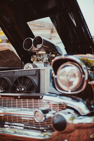 A classic car with an open hood revealing a supercharged engine. The front grille features a pair of large black fans, and the chrome details are prominent, reflecting sunlight. The image captures the mechanical intricacies and polished metal surfaces.