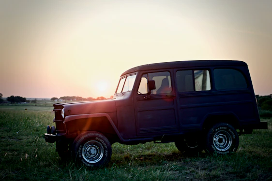 A vintage Bronco parked on a narrow mountain trail surrounded by tall pines under a golden sunset.