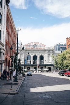 A historic train station with a large sign that reads 'Union Station Travel by Train' is at the end of a wide street lined with red brick and white buildings. There are cars parked along the street and people walking on the sidewalk. The sky is partly cloudy, and there are streetlamps and trees present.