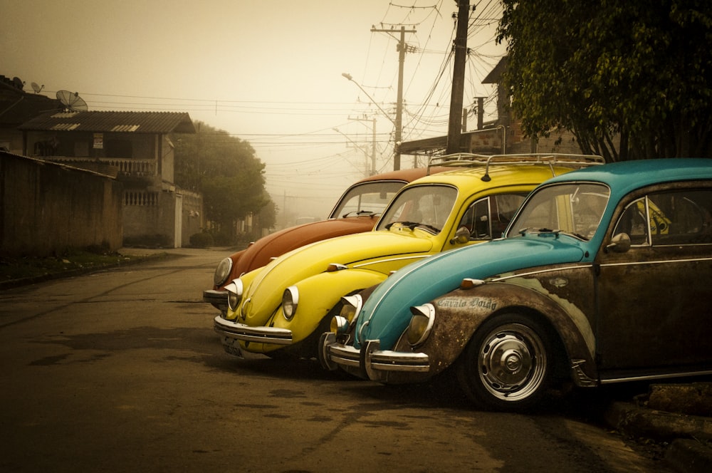 drei Volkswagen Käfer Coupés in verschiedenen Farben, die in der Nähe von Häusern geparkt sind