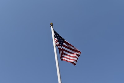 A lone American flag flutters against a clear blue sky, mounted on a tall flagpole with a gold finial.