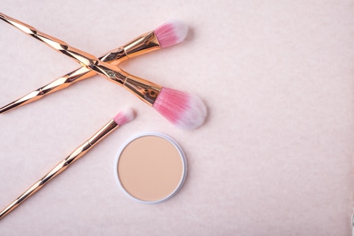 Three makeup brushes with pink bristles and rose gold handles are placed artfully on a light beige background. Next to them is a compact case of pressed powder.