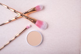 Three makeup brushes with pink bristles and rose gold handles are placed artfully on a light beige background. Next to them is a compact case of pressed powder.