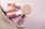 A collection of cosmetic items including a bottle of perfume, a round compact powder, a pink lipstick, a nail polish, and a makeup brush are arranged on a soft pink background. These items are partially zipped inside a beige pouch with a gold zipper.