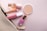 A collection of cosmetic items including a bottle of perfume, a round compact powder, a pink lipstick, a nail polish, and a makeup brush are arranged on a soft pink background. These items are partially zipped inside a beige pouch with a gold zipper.