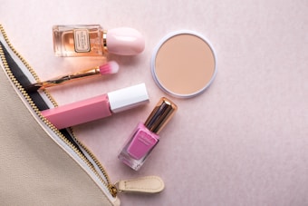 A collection of cosmetic items including a bottle of perfume, a round compact powder, a pink lipstick, a nail polish, and a makeup brush are arranged on a soft pink background. These items are partially zipped inside a beige pouch with a gold zipper.