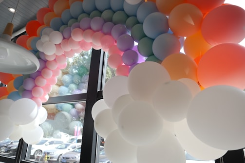 A rainbow-shaped balloon arch made of numerous balloons in pastel colors, set near large glass windows that reflect the balloons. Multiple cars are visible through the windows in the background, indicating an indoor setting with an outdoor view.
