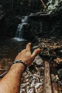 A hand wearing a colorful beaded bracelet is extended towards a small waterfall in a forest setting. The atmosphere is natural and serene, with visible rocks and tree roots around a water stream.