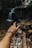 A hand wearing a colorful beaded bracelet is extended towards a small waterfall in a forest setting. The atmosphere is natural and serene, with visible rocks and tree roots around a water stream.