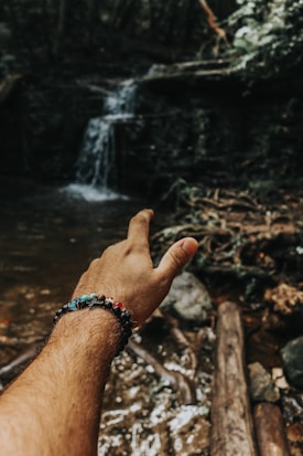 A hand wearing a colorful beaded bracelet is extended towards a small waterfall in a forest setting. The atmosphere is natural and serene, with visible rocks and tree roots around a water stream.