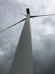 A tall wind turbine with large blades set against a cloudy sky, viewed from a low angle perspective.