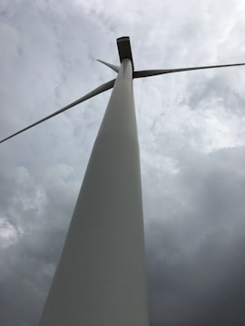 A tall wind turbine with large blades set against a cloudy sky, viewed from a low angle perspective.