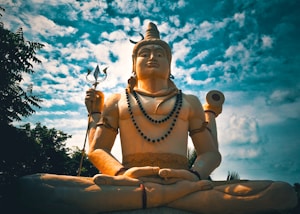 A large statue of a deity is seated in a meditative pose under a partly cloudy sky. The figure holds a trident in one hand and a drum in another, adorned with beads and ornaments. The backdrop is composed of trees and a vibrant sky, creating a serene and majestic atmosphere.