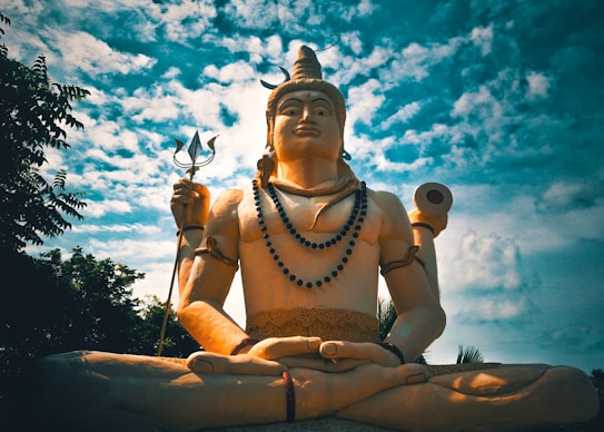 A large statue of a deity is seated in a meditative pose under a partly cloudy sky. The figure holds a trident in one hand and a drum in another, adorned with beads and ornaments. The backdrop is composed of trees and a vibrant sky, creating a serene and majestic atmosphere.