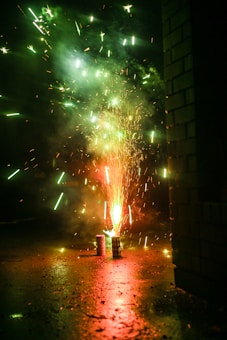 A burst of fireworks illuminates the dark surroundings with vibrant sparks and bright colors. The ground reflects the glow, and a nearby brick wall is partially visible on the right. Smoke trails and star-like sparkles fill the air, creating a lively and energetic visual display.