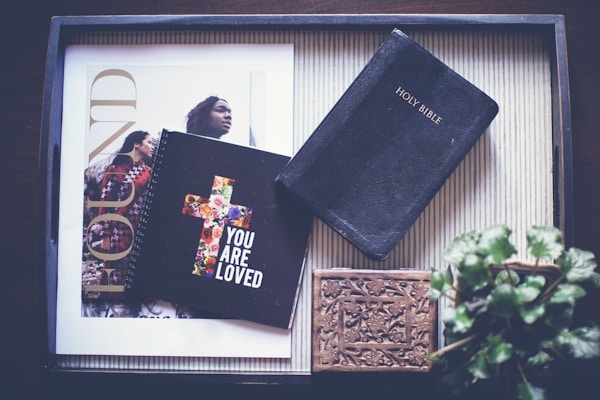 An overhead view of a tray containing a Holy Bible, a notebook with a floral cross design bearing the phrase 'YOU ARE LOVED', a decorative wooden box, and a magazine featuring a cover with two people. There is also a potted plant partially visible in the corner.