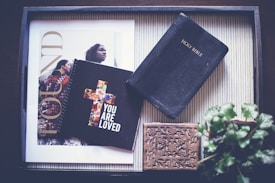 An overhead view of a tray containing a Holy Bible, a notebook with a floral cross design bearing the phrase 'YOU ARE LOVED', a decorative wooden box, and a magazine featuring a cover with two people. There is also a potted plant partially visible in the corner.