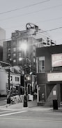 A street scene features a mix of modern and older buildings, including a high-rise with unique architectural elements. There are streetlights, multiple storefronts with signage, and a visible street sign for Spring Garden Road. Sidewalk advertising boards add to the urban atmosphere.