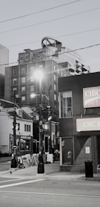 A street scene features a mix of modern and older buildings, including a high-rise with unique architectural elements. There are streetlights, multiple storefronts with signage, and a visible street sign for Spring Garden Road. Sidewalk advertising boards add to the urban atmosphere.