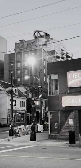 A street scene features a mix of modern and older buildings, including a high-rise with unique architectural elements. There are streetlights, multiple storefronts with signage, and a visible street sign for Spring Garden Road. Sidewalk advertising boards add to the urban atmosphere.