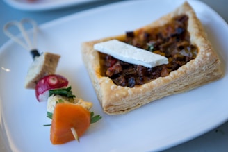 A rectangular puff pastry filled with a mixture of diced vegetables and topped with a slice of cheese is placed on a white plate. Next to it, a small skewer holds a cube of bread, a slice of radish, cilantro leaves, and a thin slice of carrot.