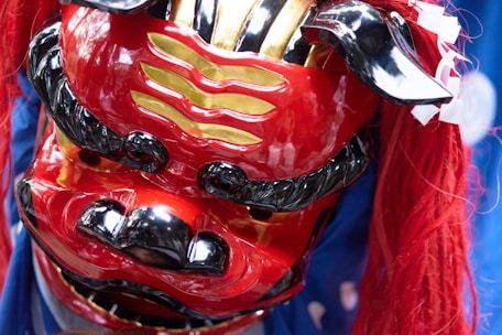 A colorful and intricately designed traditional mask featuring bold red, gold, and black colors. The mask has a fierce and expressive face with exaggerated features, including a wide mouth and large eyes. Long, vibrant red hair cascades from the top, adding to its striking appearance.