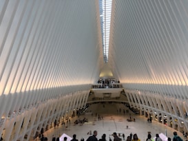 An interior space with a large, futuristic design featuring white, ribbed walls that curve upward to a skylight. People are walking and gathered in small groups on the expansive, open floor below.