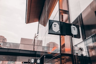 A modern storefront with reflective glass panels and an elevated walkway showing the logo of a brand featuring a stylized ape face. The architecture is sleek with an urban environment reflected in the windows, under a cloudy sky.